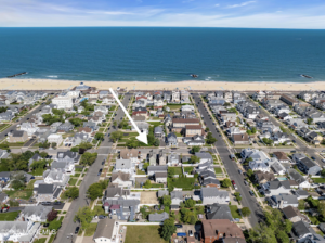 Wide aerial perspective of the sandy beach and wooden boardwalk in Belmar, New Jersey, with waves crashing on the shore and coastal houses in the background.
