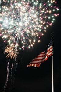 Fireworks lighting up the night sky above an American flag waving on a flagpole