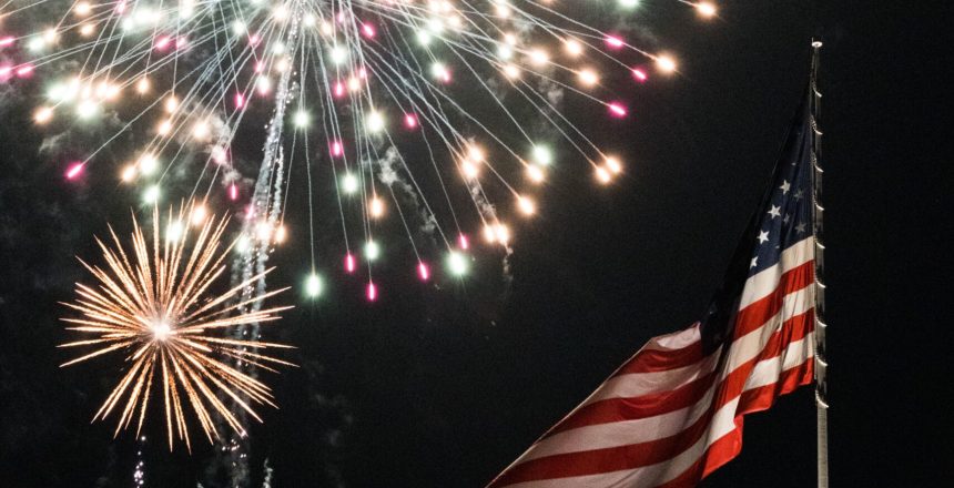 Fireworks lighting up the night sky above an American flag waving on a flagpole